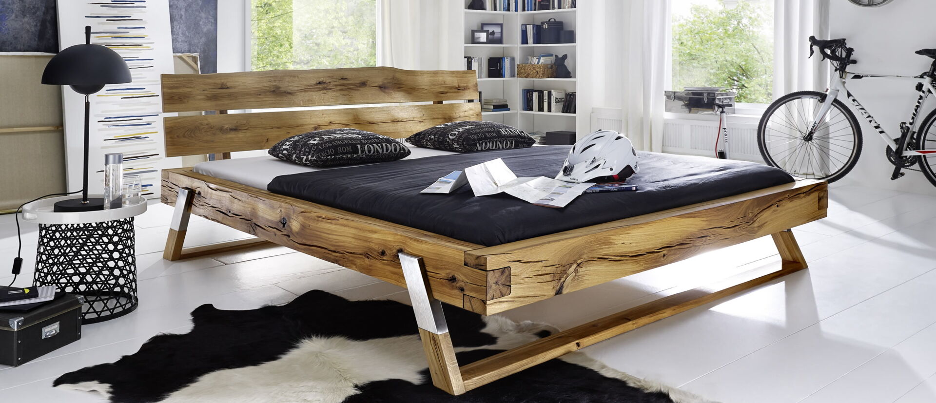 Ein robustes Holzbett mit schwarzen Kissen und Fahrrad im modernen Schlafzimmer, erhältlich bei Möbel Lutz in Obernbreit.